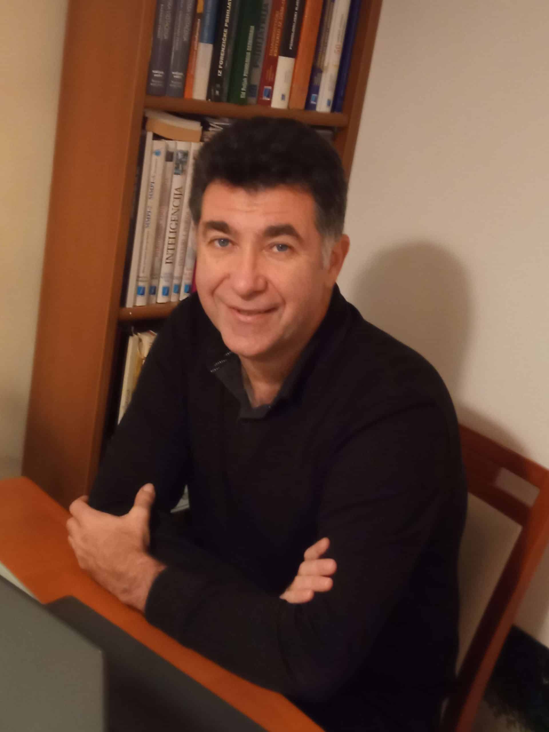 Psychologist Romel Krajačić smiling at the camera, sitting at a desk with bookshelves in the backgro.