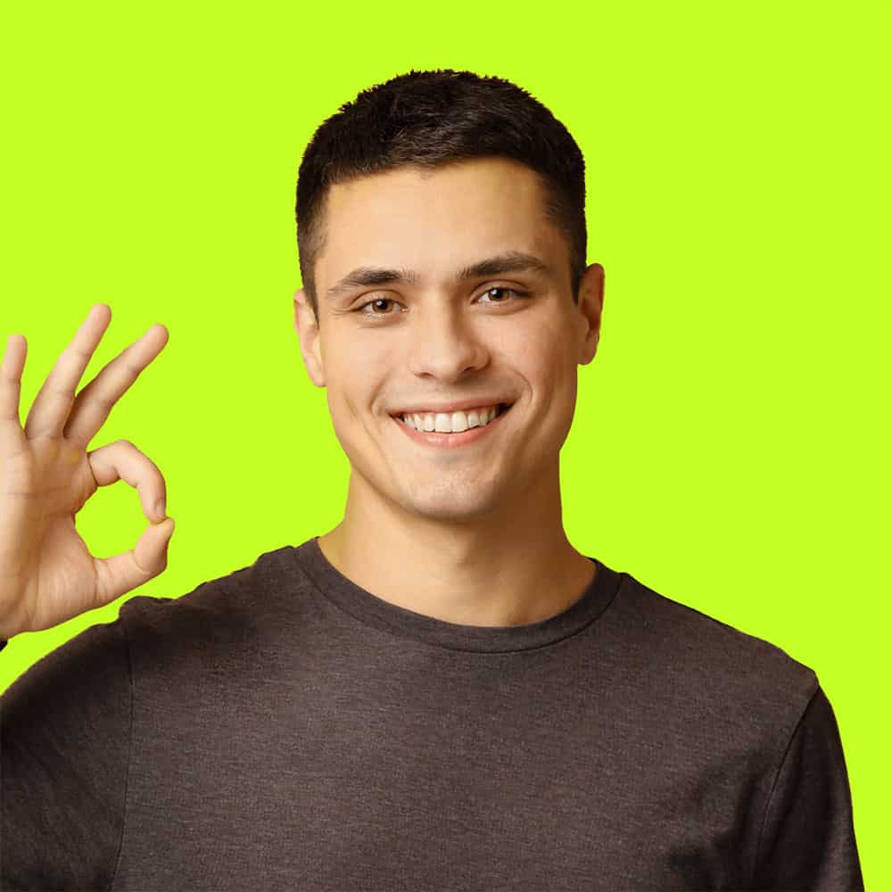 Young man making an OK sign, smiling confidently against a vibrant green backdrop.